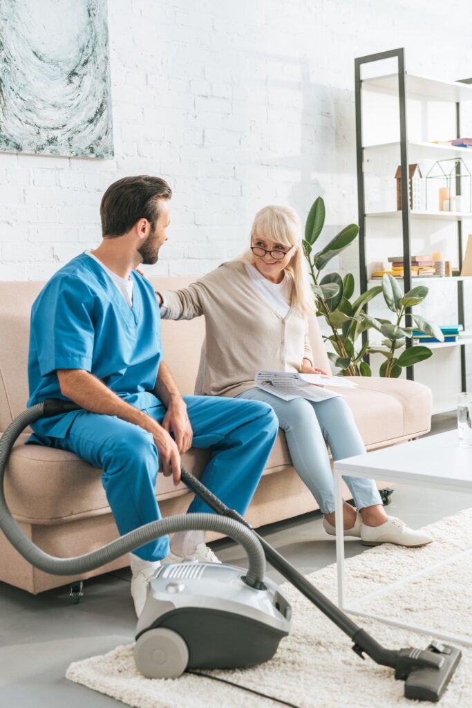 smiling senior woman sitting on couch and looking at caregiver with vacuum cleaner