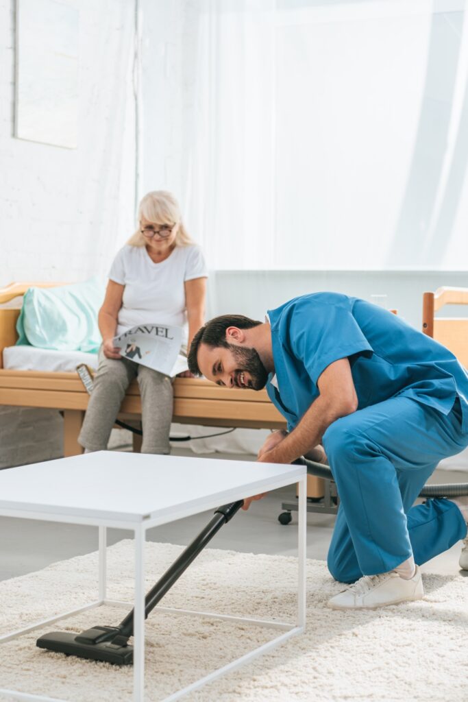 young caregiver cleaning carpet with vacuum cleaner while senior woman reading newspaper on bed